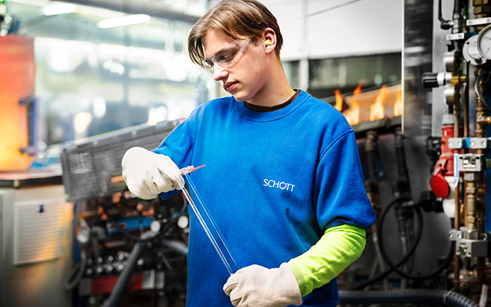 Eine Person in blauer Arbeitskleidung formt ein dünnes Glasrohr an einer Produktionsmaschine.