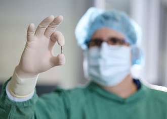Ein Arzt in OP-Kleidung hält den winzigen Sensor von Kitea Health zwischen Daumen und Zeigefinger. 