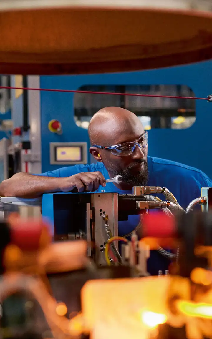 Junger Mann im SCHOTT T-Shirt prüft mit einem Schraubenschlüssel in der Hand eine Maschine, aus der Flammen kommen und Glas angeschmolzen wird