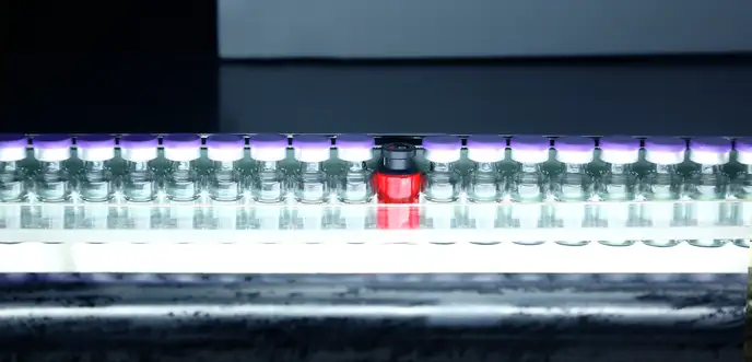 Vials with purple caps on a production line under a light, with one red vial standing out among them.