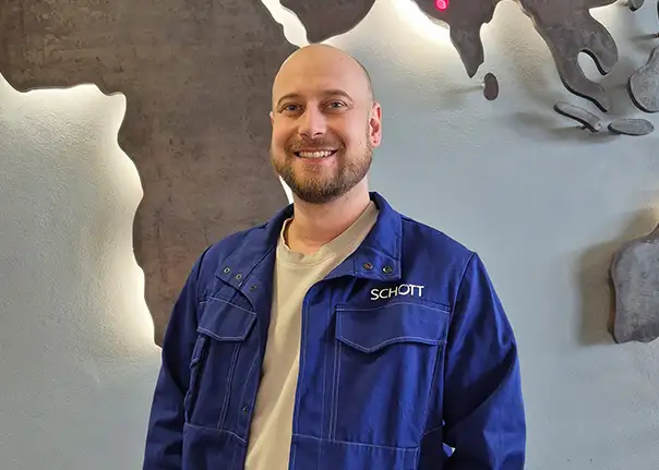 Eine Person in blauer SCHOTT‑Arbeitskleidung steht vor einer Wand.