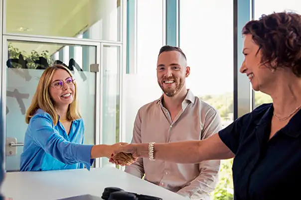 Drei Personen sitzen an einem Besprechungstisch, während zwei von ihnen sich zur Begrüßung die Hand geben.