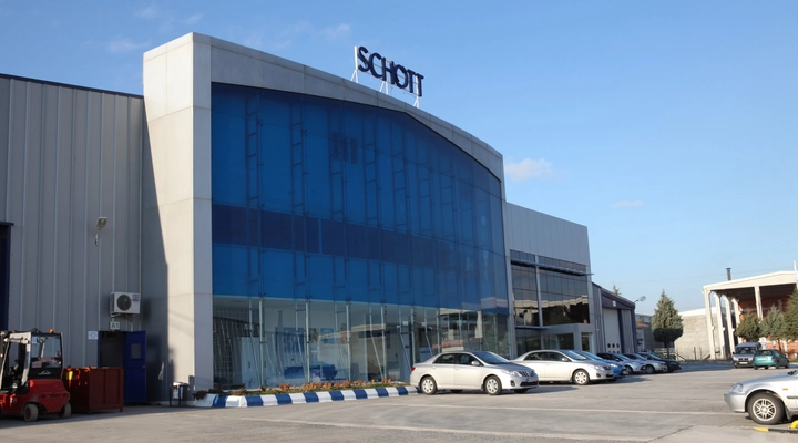Exterior view of a modern SCHOTT industrial building with blue glass facade, company logo on top, and parked cars in front