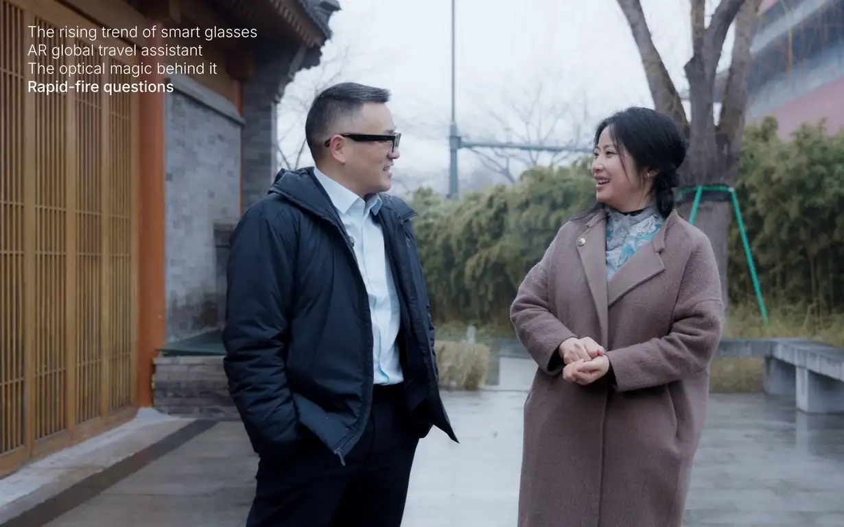 Two people standing and talking outdoors near a traditional-style building on a cloudy day, with trees and a paved walkway in the backdrop.
