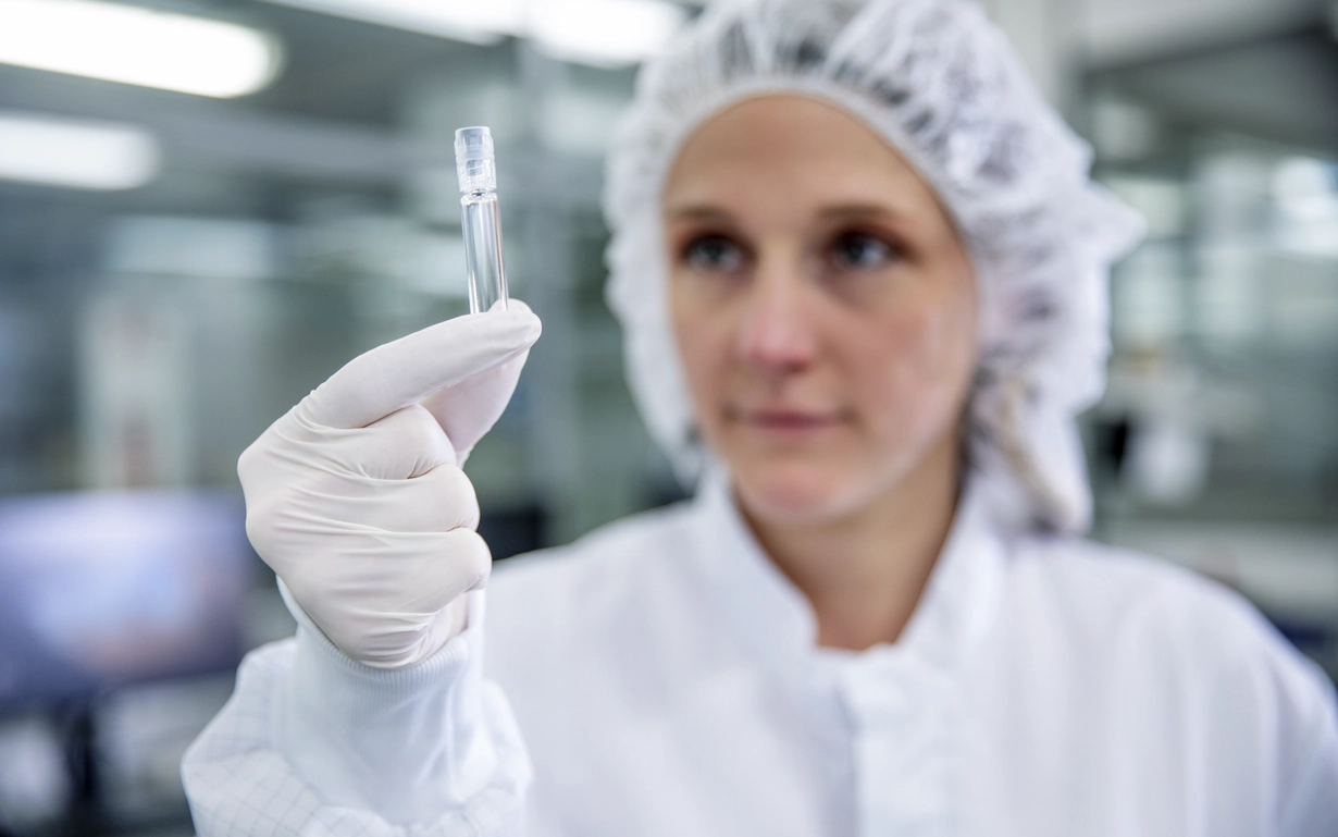 Woman in cleanroom clothing holds up a prefillable glass syringe.