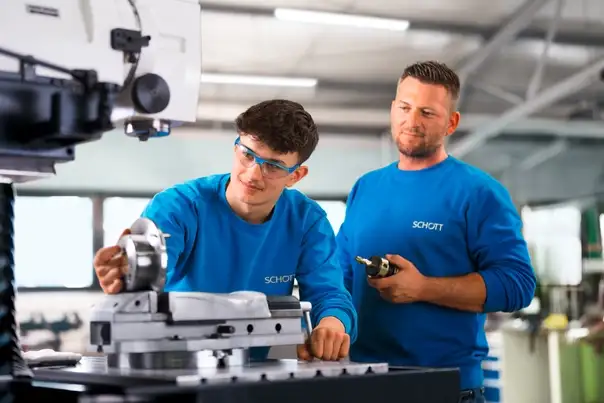 Zwei Personen in blauen Arbeitsshirts arbeiten gemeinsam an einer Fräsmaschine und justieren ein Metallbauteil.
