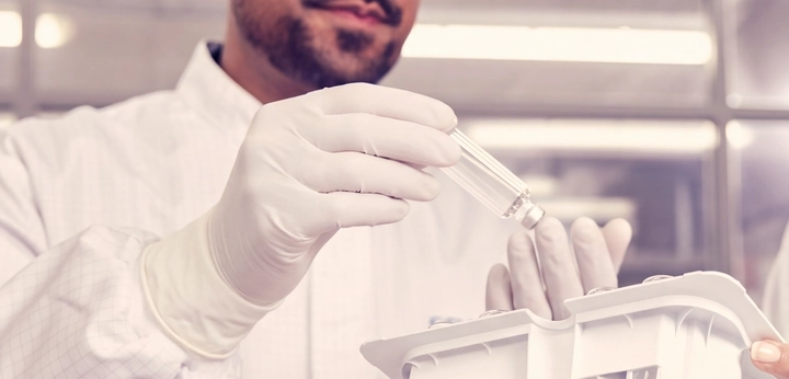 Two lab technicians in cleanroom suits inspect a cartridge tray of small glass vials in a laboratory.